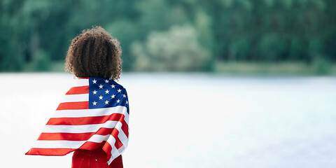 Young African american woman at nature background with an American USA flag on her shoulders. USA...