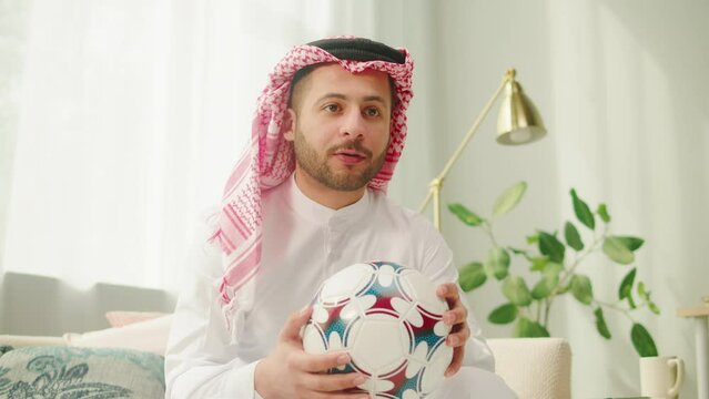 Middle eastern football fan supporting favorite team, watching match on TV. Young man holding soccer ball. Wearing traditional Islamic male clothes. Worship and culture concept.
