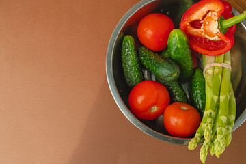Vegetables lie in a metal bowl: tomatoes, asparagus, cucumbers, red bell peppers. on a wooden board and brown background. place for text. view from above