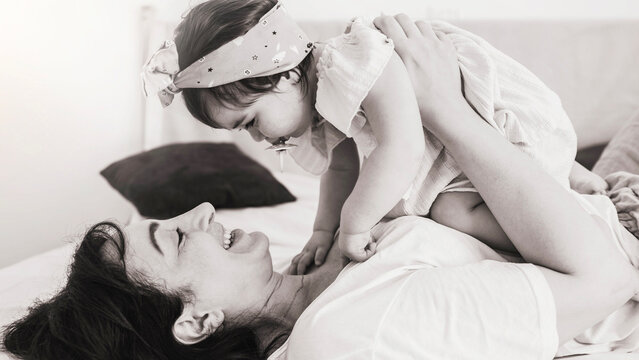 Portrait of beautiful mother playing with her kid at home - Happy mom having fun holding her baby on the bed - Black and white filter