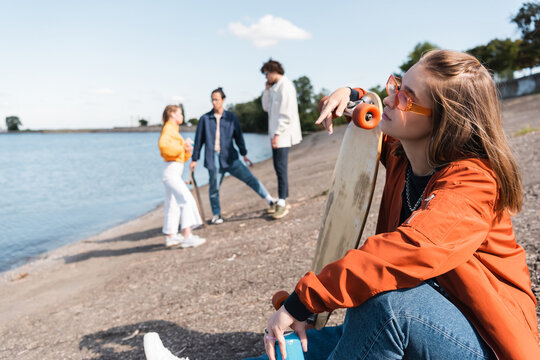 young woman in eyeglasses sitting on river bank with penny board near friends on blurred background.