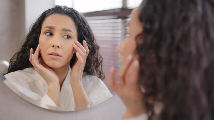 Upset worried young hispanic woman standing in bathroom looking in mirror worries about first wrinkles sad girl unhappy with condition of skin on face frustrated bad skincare product feeling stress