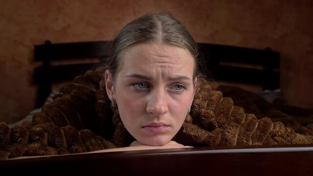 Young girl is sad lying on a bed under a blanket close-up. he face of a beautiful girl looks sadly and dejectedly at one point, as if she is about to cry. Disorder.