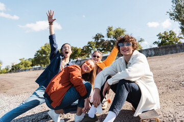 excited asian woman with raised hand screaming near smiling friends outdoors.