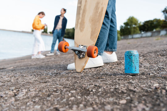 partial view of skater with longboard near soda can and blurred friends on river bank.