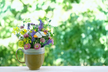 Beautiful meadow flowers and butterfly in cup on wooden table in garden, green natural abstract background. Summer season. floral composition in rustic style. copy space