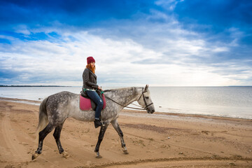 wearing jeans, jacket and spring hat horsewoman takes horse along coast