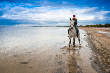 wearing jeans, jacket and fall hat woman rides a gray horse along the ocean