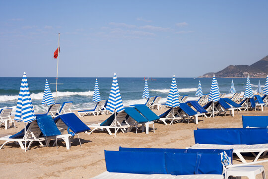 Beach with closed striped umbrellas and deck chairs on the background of the sea. An equipped beach is waiting for guests to relax. White and blue stripes. Mediterranean Sea - Powered by Adobe