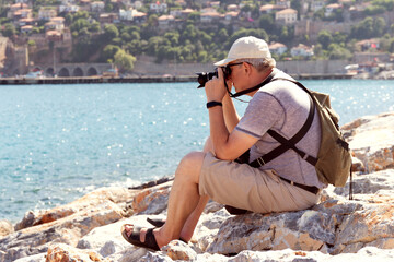 Man with camera sitting on the rocks. Tourist taking photo of sea against maritime city. Blurred background. Selective focus.