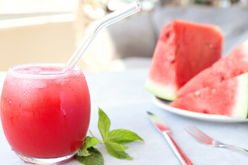 Close-up of cold tasty watermelon smoothie in the glass with straw, mint and watermelon slices on plate on white table. Blurred background. Selective focus.