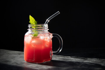 Close-up of fresh watermelon smoothie with ice and mint in jar with straw on black background. Dark low key photo. Selective focus. Blurred background.