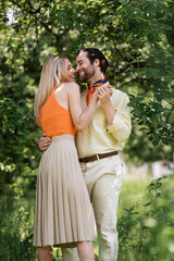 Fototapeta premium Side view of cheerful stylish couple holding hands near trees in summer park.