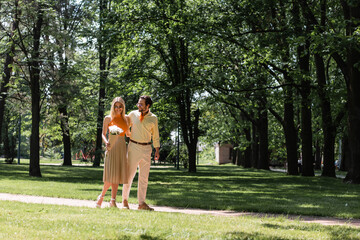 Fototapeta premium Smiling woman in sunglasses holding bouquet near boyfriend in summer park.