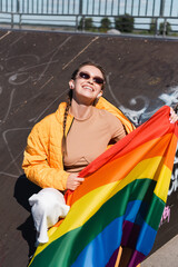 happy woman in wireless earphones sitting with lgbt flag on ramp in skate park.