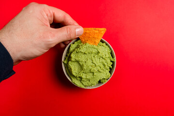 Man's hand dipping a triangle of corn in guacamole inside a ceramic bowl on a round slate plate, red background