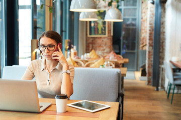 Serious multitasking young lady with fashionable wristwatch sitting at table in restaurant and...