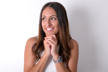 Fototapeta premium Happy young beautiful caucasian woman wearing white Top over white background, anticipates something awesome happen, looks happily aside, keeps hands together near face, has glad expression.