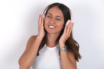 Pleased young beautiful caucasian woman wearing white Top over white background, enjoys listening pleasant melody keeps hands on stereo headphones closes eyes. Spending free time with music