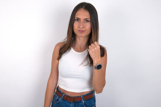 young beautiful caucasian woman wearing white Top over white background, shows fist has annoyed face expression going to revenge or threaten someone makes serious look. I will show you who is boss