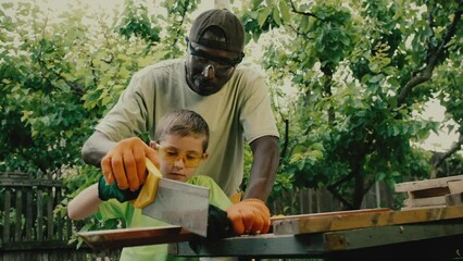 African American carpenter teaches white boy to use handsaw for woodwork. Focused schooler in goggles cuts long wooden board under control of black specialist