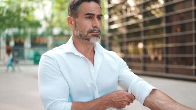 Clouse-up, mature businessman with neat beard, straightens his white shirt, walks on the street of modern city. Successful man relaxed, enjoying life