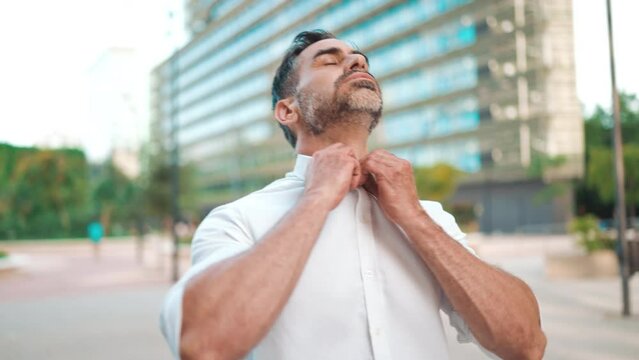 Mature businessman with neat beard, unbuttoning white shirt stands on the street of modern city. Successful man is relaxed, enjoys life. Camera moves around