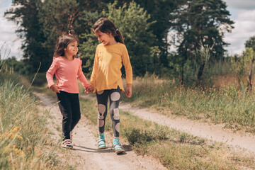 Little girl friends walk holding hands along a rural path near the forest, summer vacation