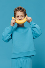  handsome, happy boy stands in blue clothes on a blue background and holds a banana in his hand, substituting it as a smile to his face. Studio photo with empty space for advertising insert
