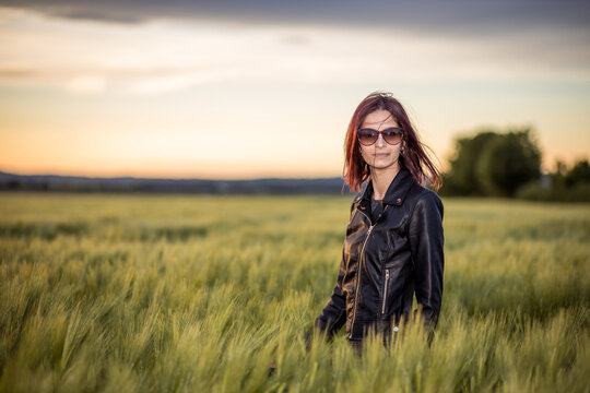 Young Caucasian woman standing among long green grass in the field against dusk sky at sunset