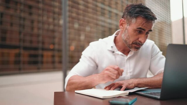 Mature businessman with beard, user on laptop pc computer and make notes sit at cafe outdoors. Successful man sitting at cafe table outdoors on nature Mobile office freelance