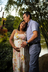 man with closed eyes hugging and touching belly of pregnant wife with lake and tropical nature in the background and blue sky
