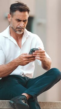 Mature businessman with neat beard uses mobile phone sits on bench in the financial district in the city. Successful man scrolls through information on smartphone. Camera zoom