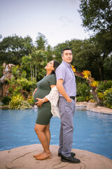husband and pregnant wife touching each other's backs near swimming pool surrounded by tropical jungle