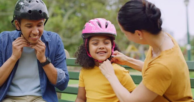 A happy diverse family fitting a pink helmet on their daughter while sitting on a park bench before having fun rollerblading outdoors. Affectionate mother and father checking their kids safety gear