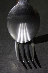 Los dientes del tenedor en luz oblicua se reflejan en la cuchara en un desenfoque, proyectando una sombra en el suelo creando un diseño abstracto original con un fondo negro