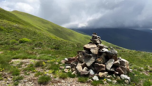 closeup of pyramid of stacked stones used for hiking trail tourist guidance with beautiful sunny mountain slope range covered in green grass in the background and cloudy sky
