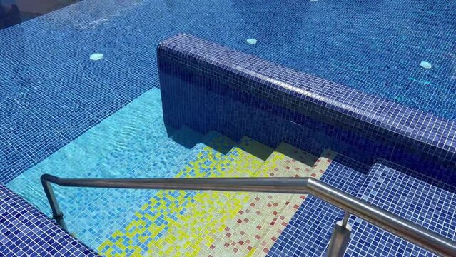 perspective view closeup of grab bars chrome ladder railings going in a outdoor swimming pool decorated with blue ceramic tiles