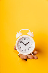 Hand holds a white alarm clock through a paper hole in yellow background. Vetical image
