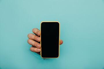 A male hand sticking out of a hole from a blue background, holds a smartphone with a black screen