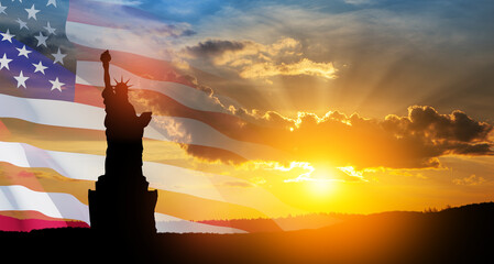 Statue of Liberty with a large american flag and sunset sky on background.