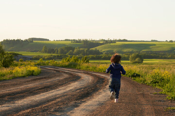 The child runs away into the distance along a dirt winding road passing through a green pasture. The soft light of the setting sun.