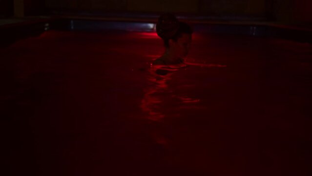 Beautiful girl floats in swimming pool in evening, lighten by red illumination. Young woman looks around fearfully while standing in water at night. Fright, surprise, risk, horror, adventure