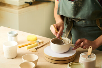 Close-up of girl painting clay mug with glaze. Woman coloring pottery in workshop with a paintbrush. Painter in green apron glazing clay pot.