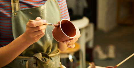 Close-up of girl painting clay mug with glaze. Woman coloring pottery in workshop with a paintbrush. Painter in green apron glazing clay pot. - 513057659