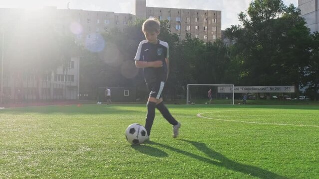 SLOW MOTION - Boy trains professionally on a football field with a ball. Training with a ball on football pitch while sunset. Active child makes a feint with football on a green field. Sport concept