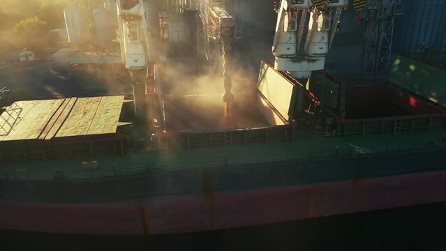 Aerial of wheat loading to bulker ship cargo hold at sea grain elevator in sea port. Wheat shipment from silo to bulk vessel via trunk at sea port terminal. Transportation of agricultural commodities.