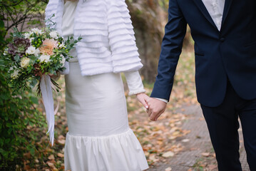 A couple of newlyweds hold hands on a walk through an autumn park. In the hands of the bride is a stylish modern bouquet of succulents, carnations and hydrangeas. Autumn view of the bride. High