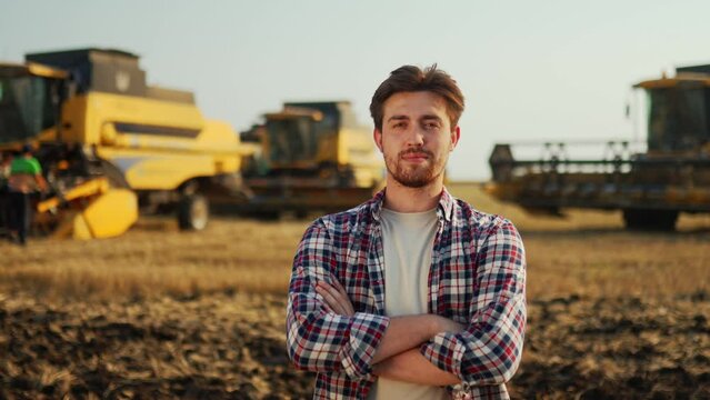 Portrait of proud farmer crossing hands on chest in harvested wheat field. Harvester machine driver standing at his combine. Smiling agronomist looking at camera. Rancher at harvesting work.