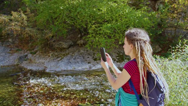 Girl tourist with backpack takes photo of beautiful natural landscape. Young woman vlogger records video for vlog on smartphone. Profile view. Female backpacker, hiker shooting landmarks on cellphone